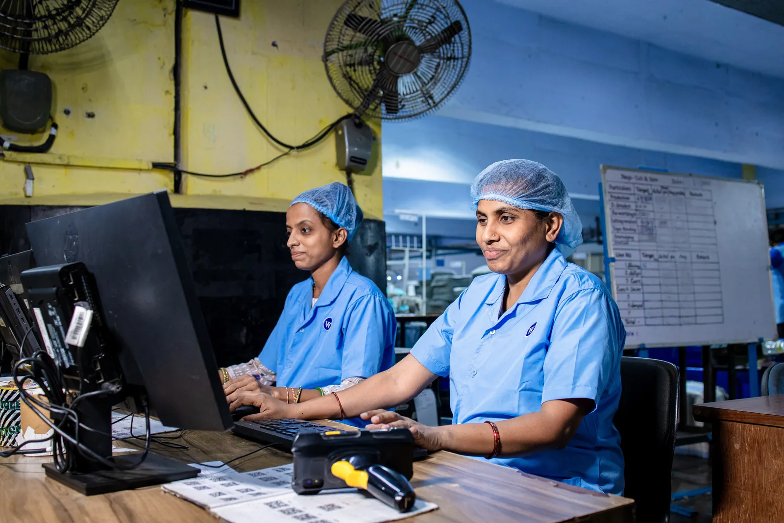 Female factory workers at desks using computers in an industrial setting, illustrating modern blue-collar employment.